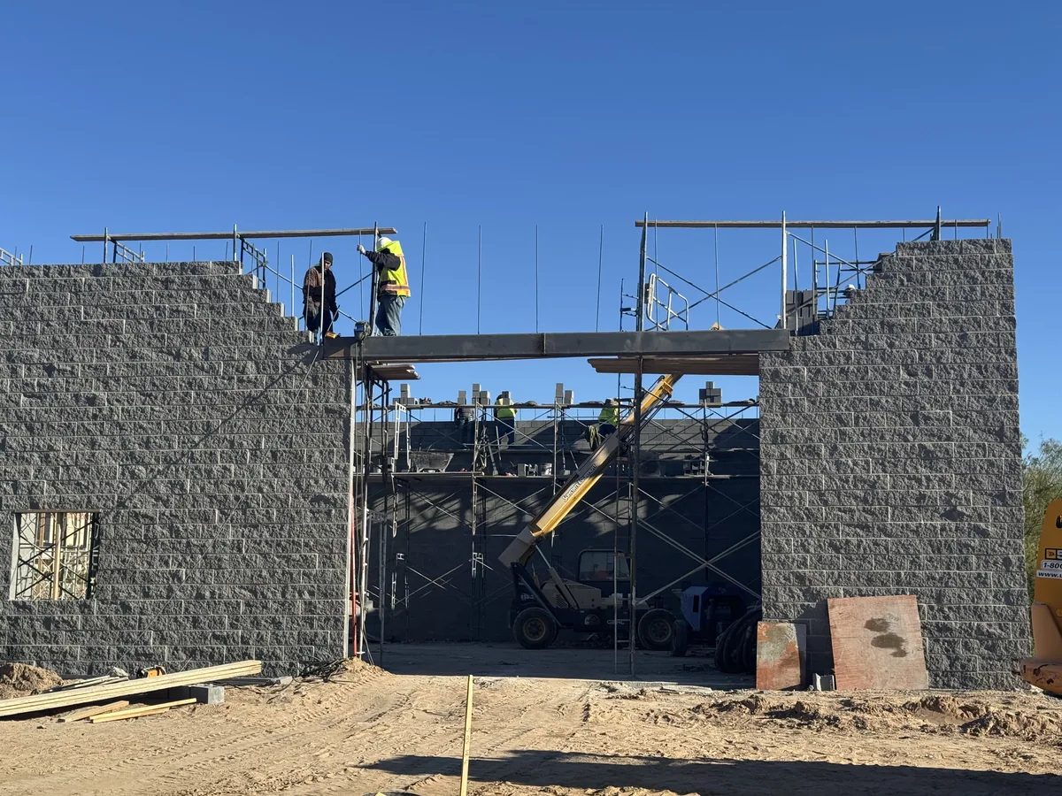 Masonry block walls of a new building under construction, with inspectors on scaffolding checking a structural steel header beam spanning the entrance opening.