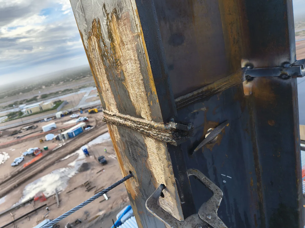 Field weld on a steel column at height, photographed during inspection above the same active construction site.